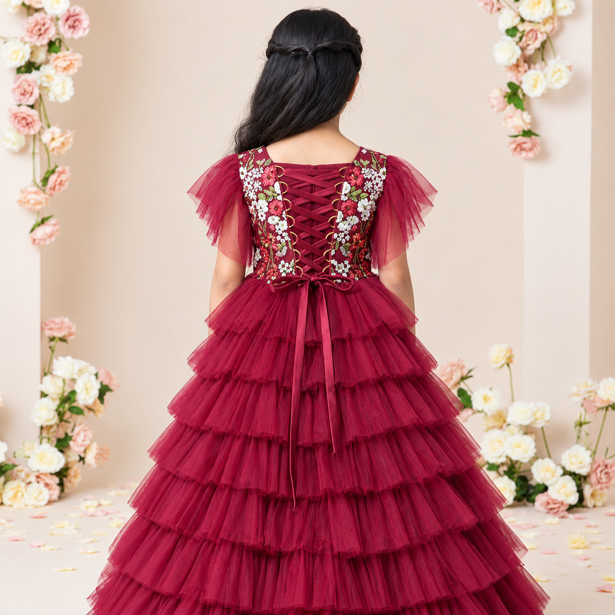 A young girl viewed from behind wearing a burgundy tulle dress with floral embroidery on the back bodice and layered ruffle skirt, standing in a decorated indoor setting with pink and white flowers.