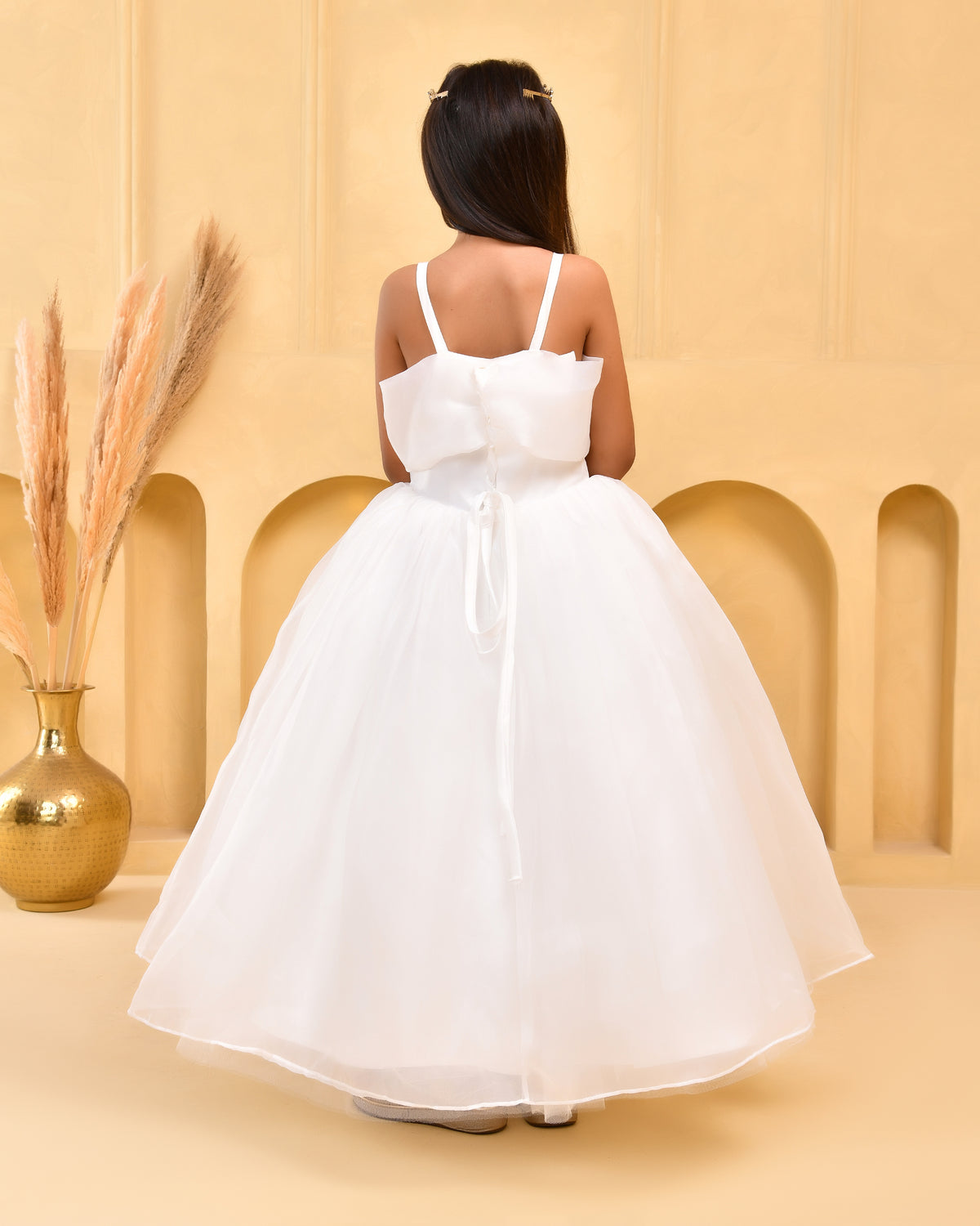 A young girl viewed from behind wearing a white sleeveless ball gown with thin straps, a large bow at the back, and a voluminous tulle skirt, standing in a warm beige studio setting with a gold vase and dried grass on the left.
