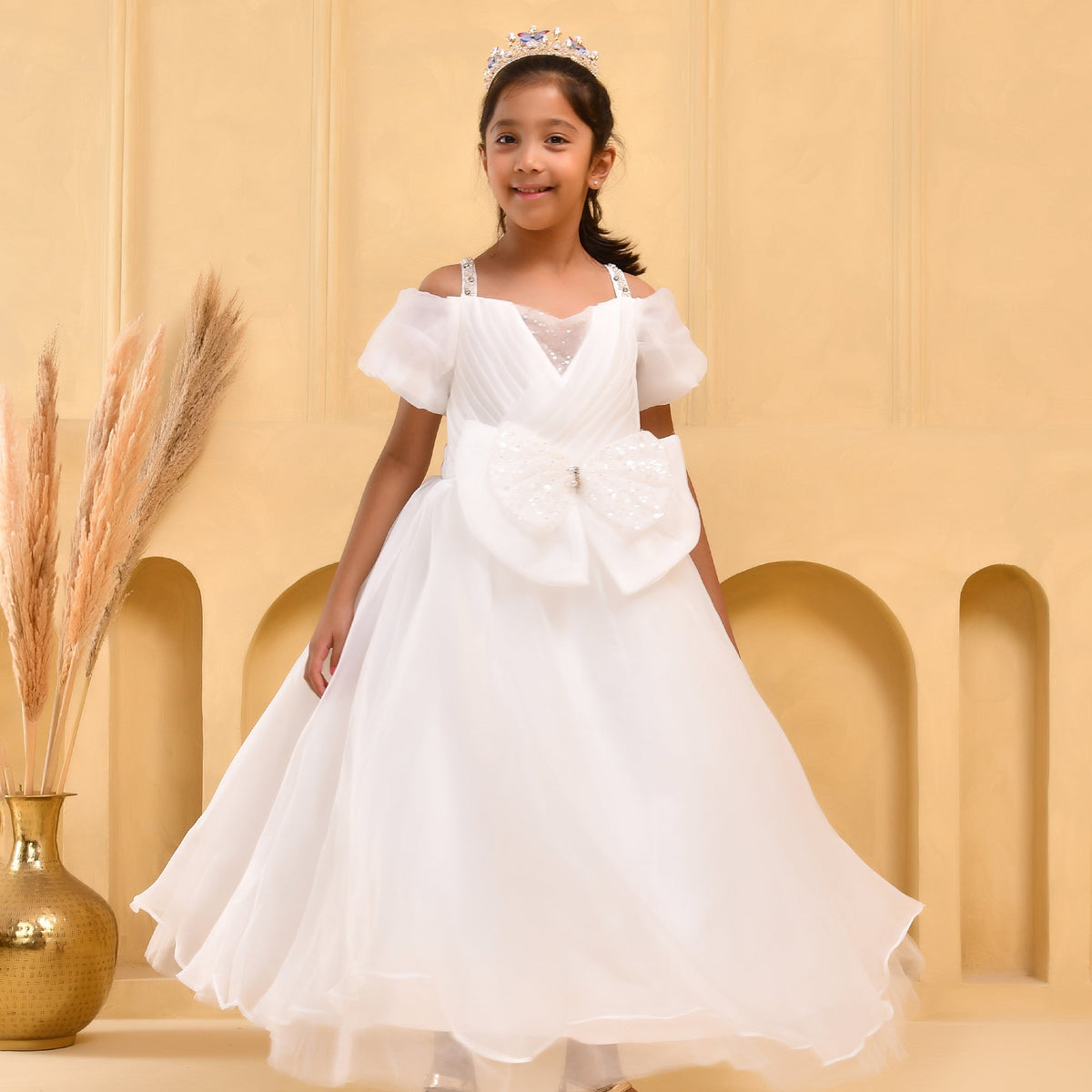 A young girl wearing a white off-the-shoulder formal dress with a large bow at the waist, standing in front of a beige arched backdrop. She is wearing a small tiara, and a gold vase with dried grass is visible on the left.