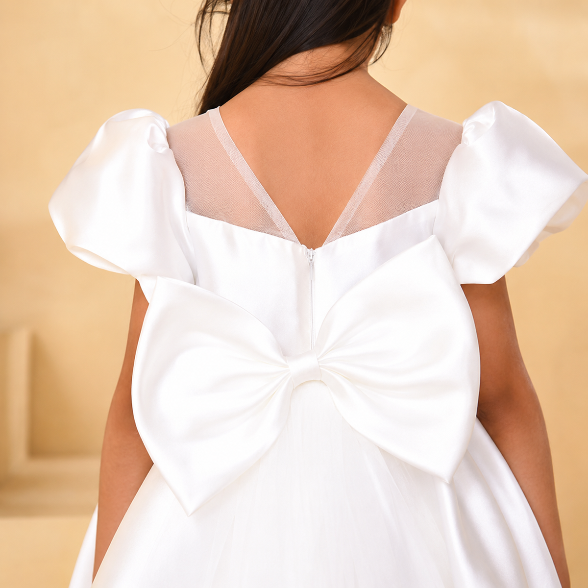 A close-up back view of a child wearing a white dress with a large bow on the back, sheer V-neck straps, and puffed short sleeves, against a warm beige background.