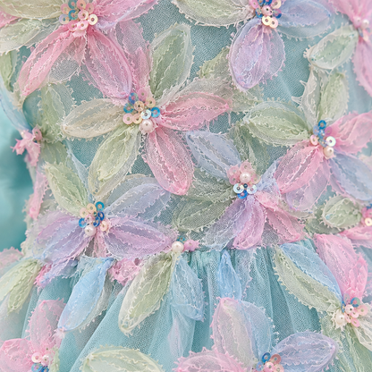 Close-up of a pastel floral fabric detail with layered tulle petals in pink, blue, and mint green, decorated with small pearl-like beads and sequins.