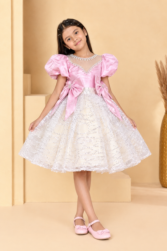 A young girl modeling a pink and white princess-style dress with puff sleeves, a lace skirt, and matching pink shoes against a warm beige studio backdrop.