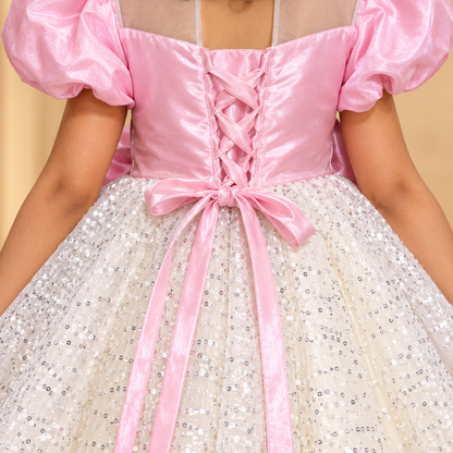 A close-up back view of a child wearing a pink and white sequined ball gown costume. The dress features a shiny pink satin bodice with puff sleeves, a lace-up back, and a voluminous white tulle skirt covered in silver sequins.