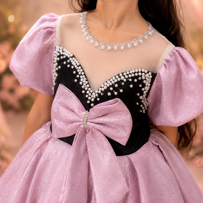 A close-up of a girl wearing a formal ball gown with shimmering light pink puff sleeves, a black sweetheart bodice decorated with white pearls, and a large matching pink bow at the waist.