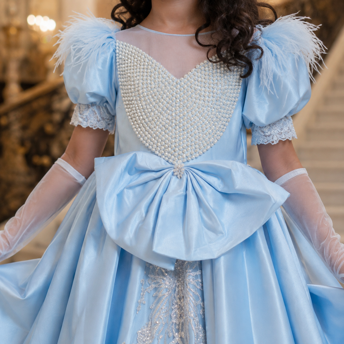 A close-up of a girl wearing a light blue ballgown costume with feathered puff sleeves, a pearl-embellished sweetheart bodice, sheer long gloves, and a large bow at the waist, photographed indoors on a staircase.