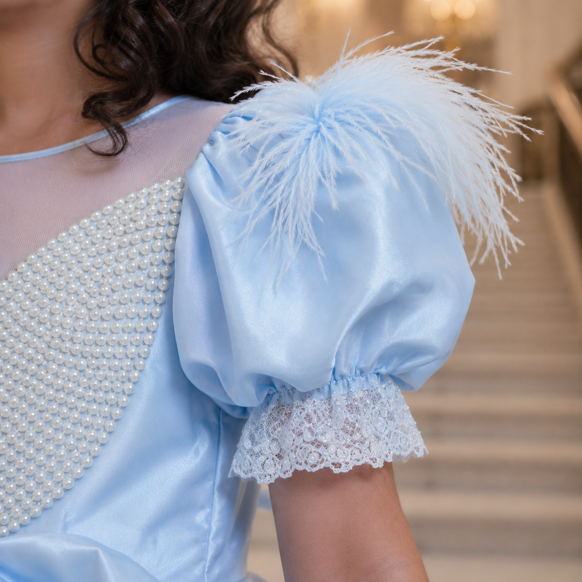 A close-up of a light blue satin dress with a large feathered puff sleeve, lace ruffle cuff, and pearl-beaded shoulder detail, worn by a model in an indoor setting.