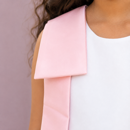 A close-up of a model wearing a light pink satin bow draped over a white sleeveless dress, set against a solid dusty pink background.