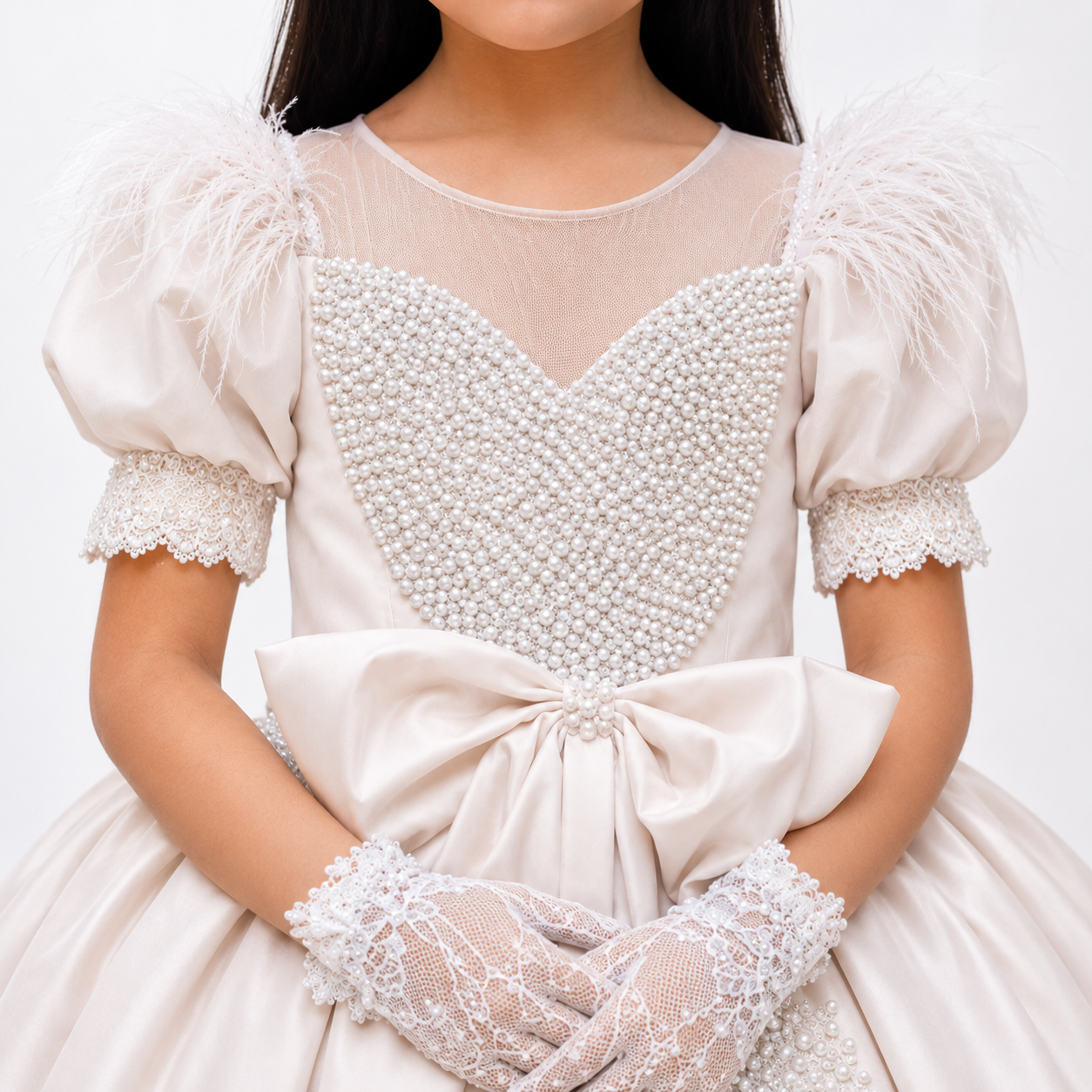 A close-up of a child wearing a formal cream or white dress with feathered puff sleeves, a beaded heart-shaped bodice, lace cuffs, and matching lace gloves against a plain light background.