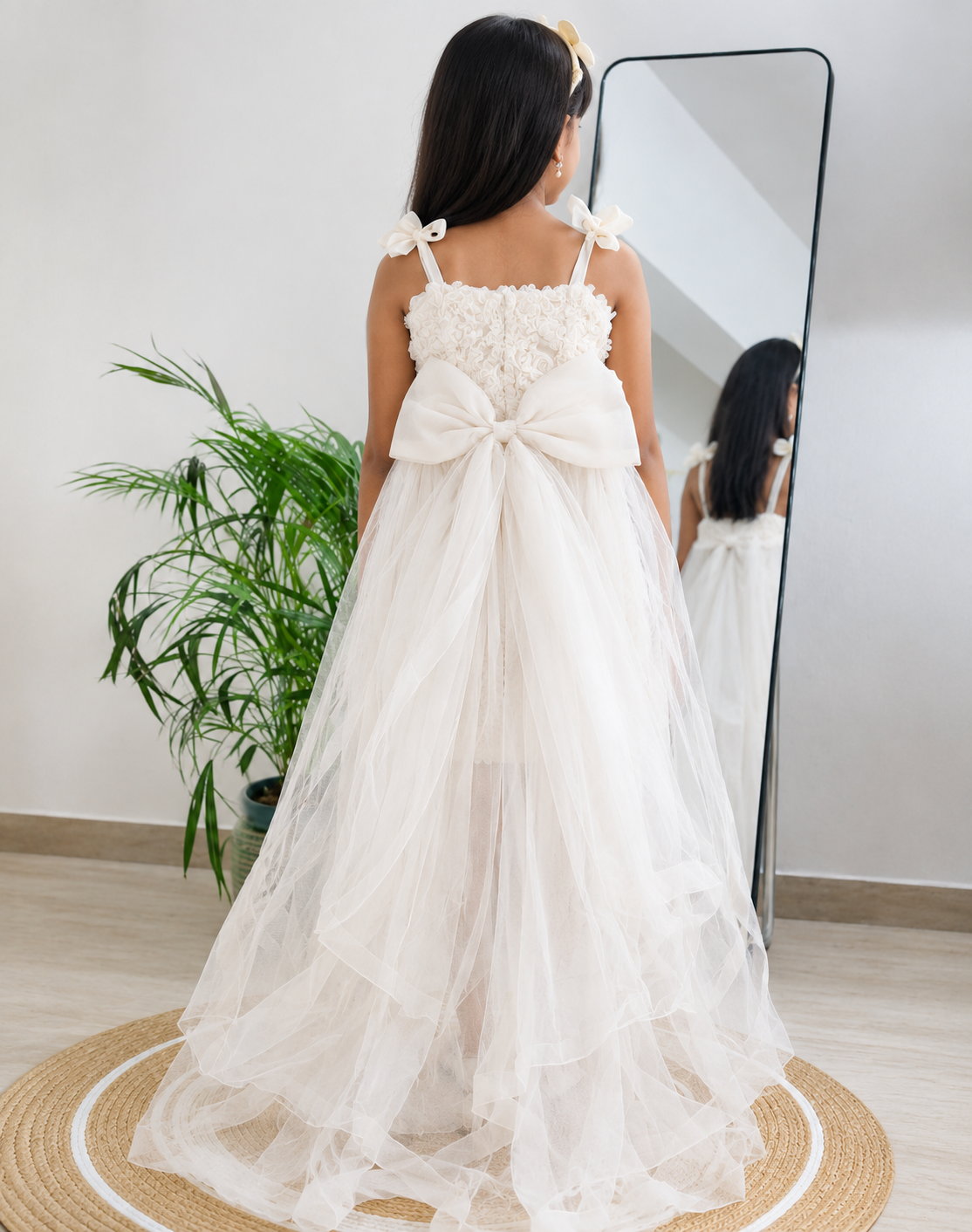 A child wearing a long white tulle dress with a large bow at the back, photographed indoors from behind near a full-length mirror and potted plant.