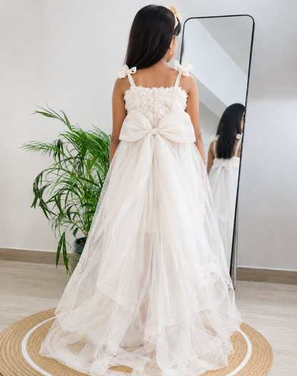 A child wearing a long white tulle dress with a large bow at the back, photographed indoors from behind near a full-length mirror and potted plant.