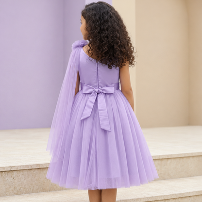 A young girl with curly dark hair is shown from behind wearing a lavender purple tulle dress with a cape-style back overlay and matching bow, standing on stone steps against a soft beige and lavender background.