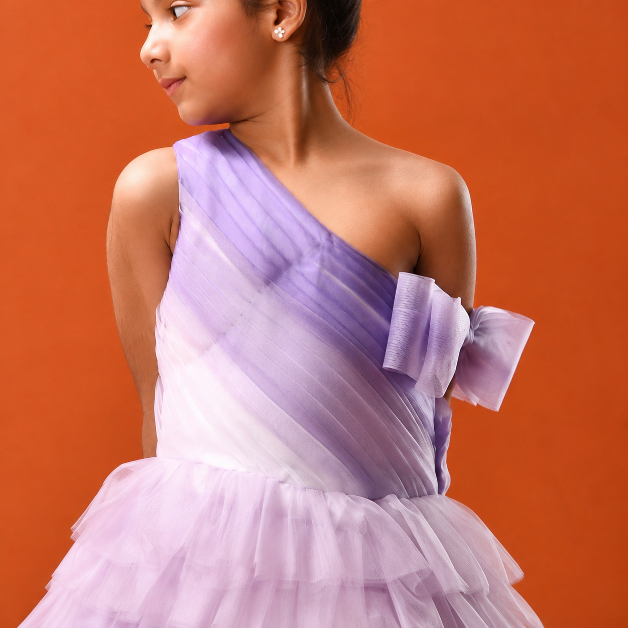 A young girl wearing a one-shoulder lavender and white tulle dress with a large bow detail on the back, posed against a solid orange background.