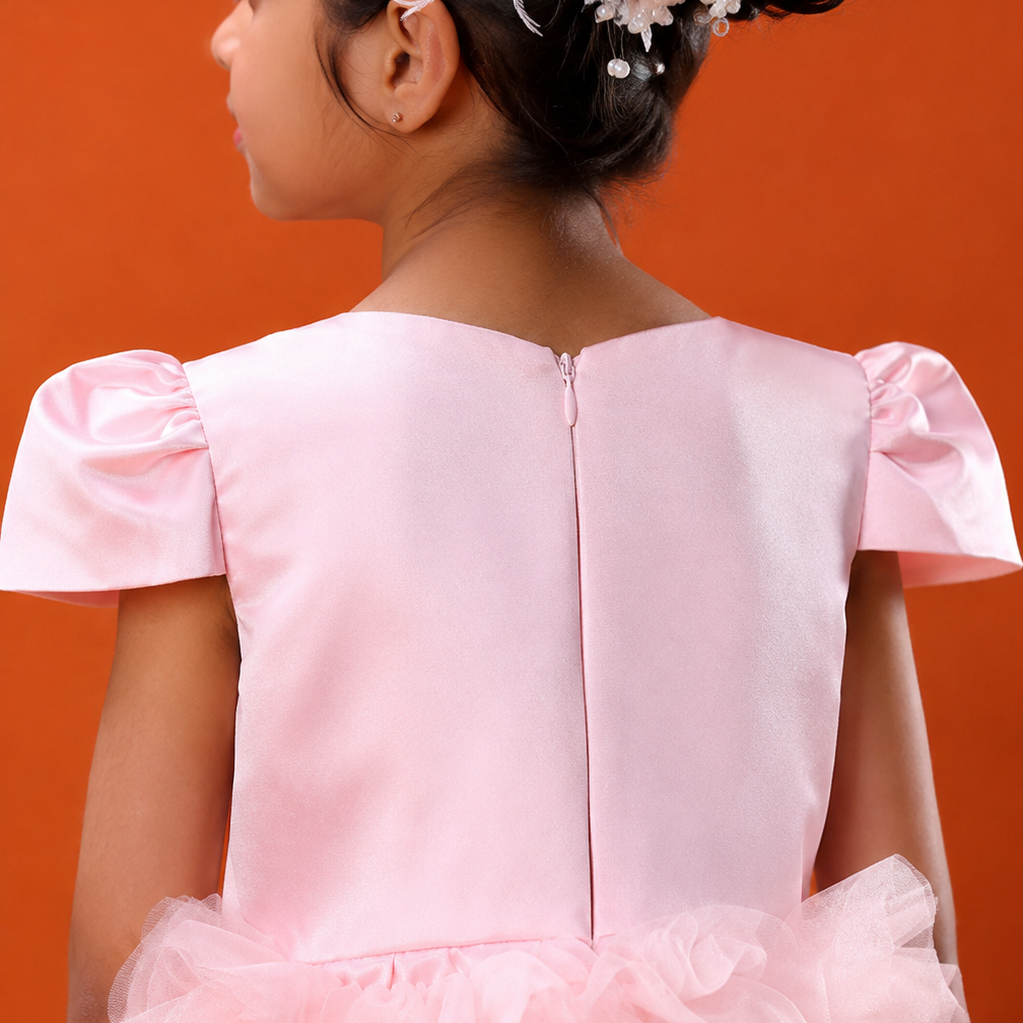 A young girl viewed from the back wearing a light pink formal dress with a smooth satin-like back panel, short flutter sleeves, and a layered ruffled tulle skirt. She is posed against a solid orange background.