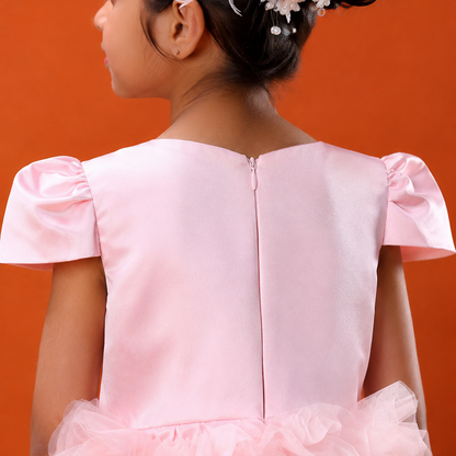 A young girl viewed from the back wearing a light pink formal dress with a smooth satin-like back panel, short flutter sleeves, and a layered ruffled tulle skirt. She is posed against a solid orange background.