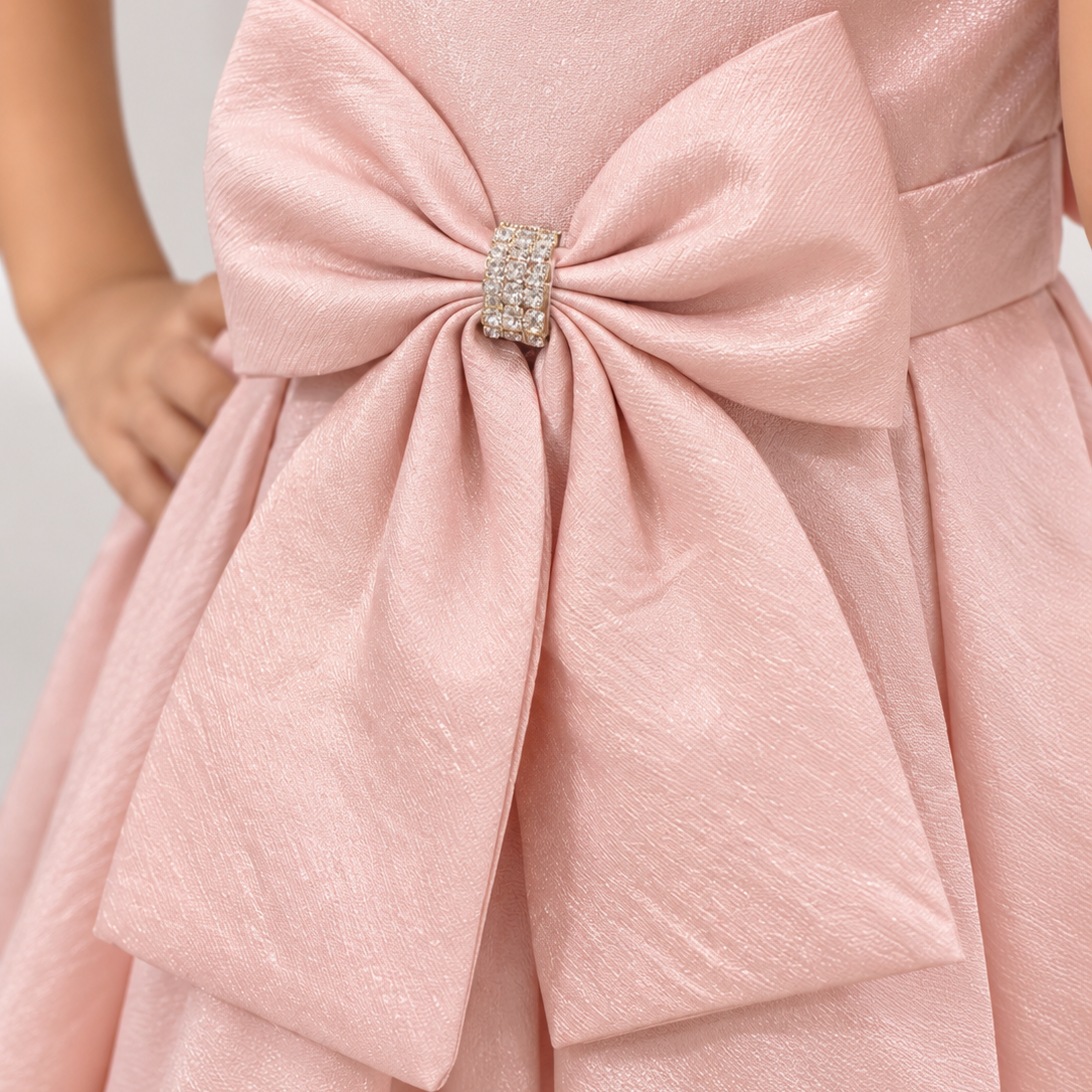 A close-up of a child wearing a metallic pink dress with a large bow at the waist secured by a rhinestone-embellished center clasp.