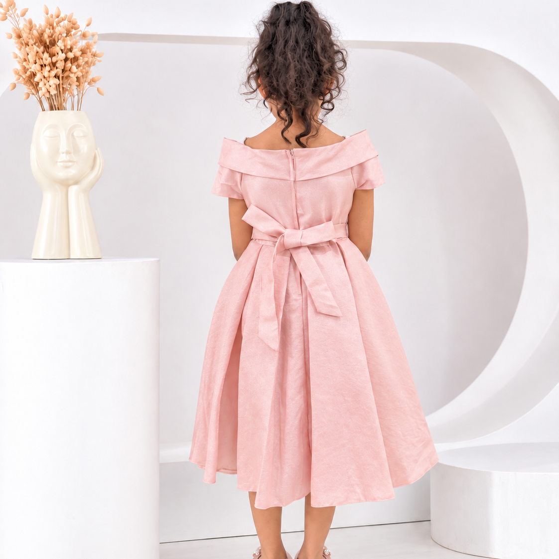 A young girl with curly dark hair is shown from behind wearing a light pink off-the-shoulder midi dress with a matching waist tie, standing in a modern white studio setting.