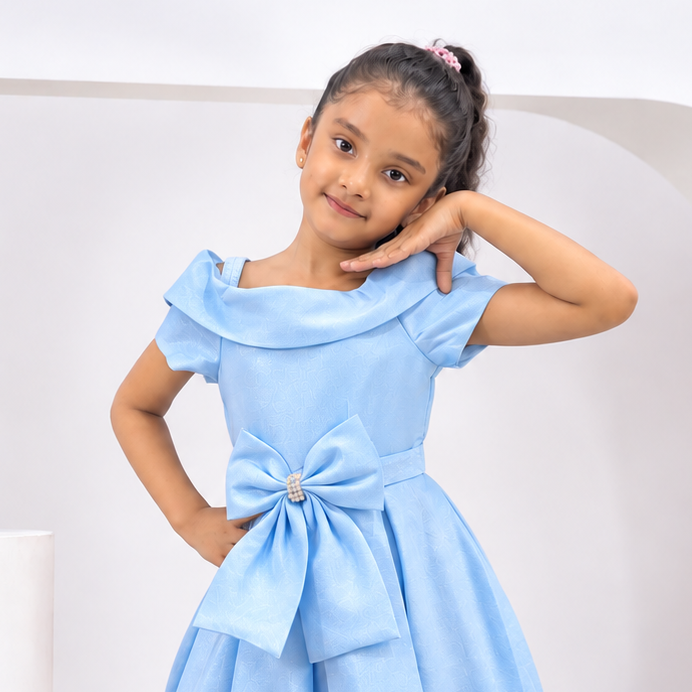 A young girl modeling a light blue midi dress with a large bow at the waist, standing in a clean white studio setting with decorative pedestals and a vase.