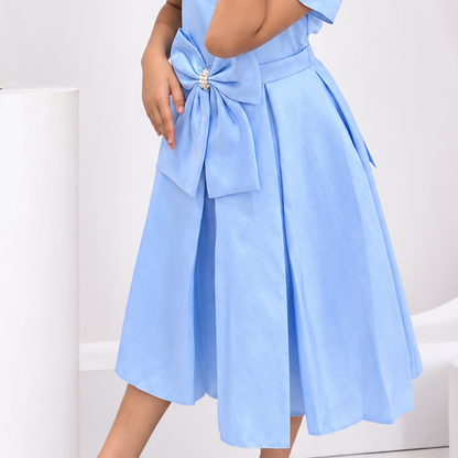 A young girl modeling a light blue midi dress with a large bow at the waist and matching blue shoes, posed in a modern white studio setting with decorative pedestals and a vase.