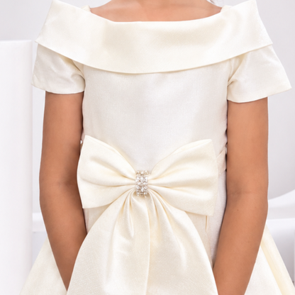 A young girl with dark curly hair wearing a cream off-the-shoulder formal dress with a large bow at the waist, standing in a modern white studio setting.