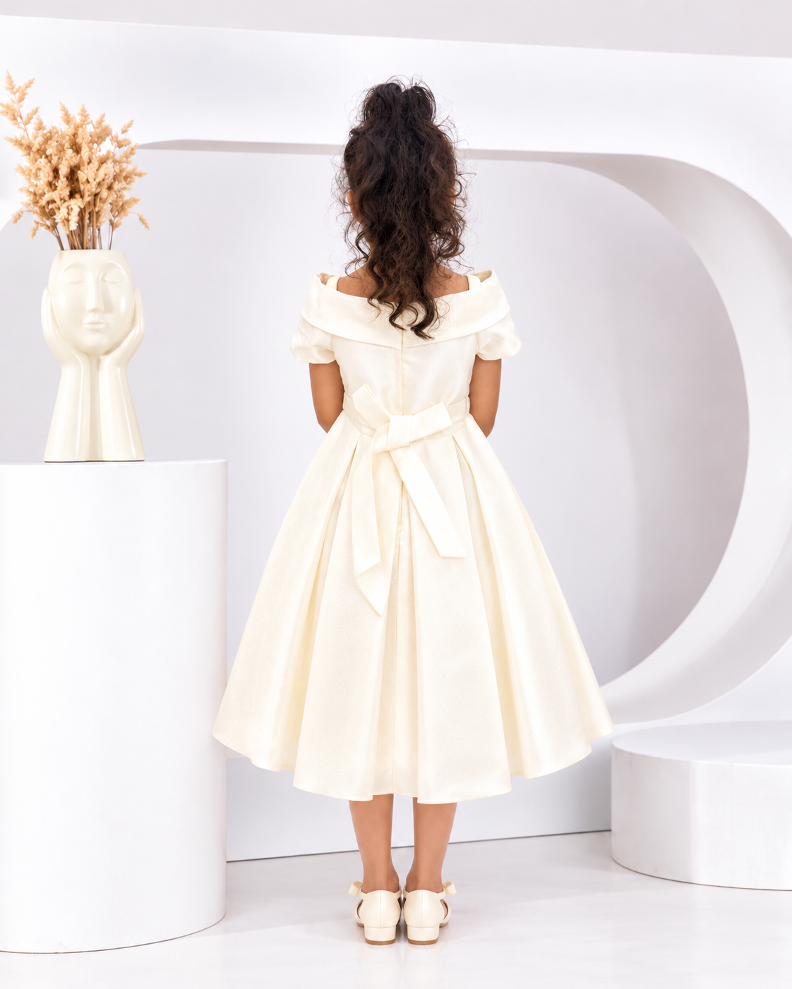 A child model viewed from behind wearing an off-the-shoulder cream-colored midi dress with a matching waist tie, standing in a minimalist white studio setting.