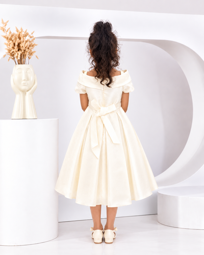 A child model viewed from behind wearing an off-the-shoulder cream-colored midi dress with a matching waist tie, standing in a minimalist white studio setting.