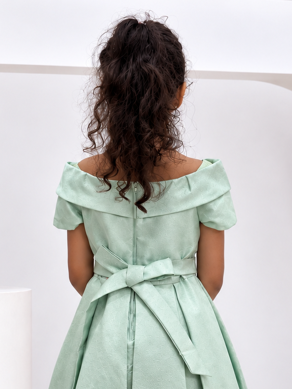 A young girl viewed from behind wearing a mint green off-the-shoulder midi dress with a matching waist tie, standing in a modern white studio setting.