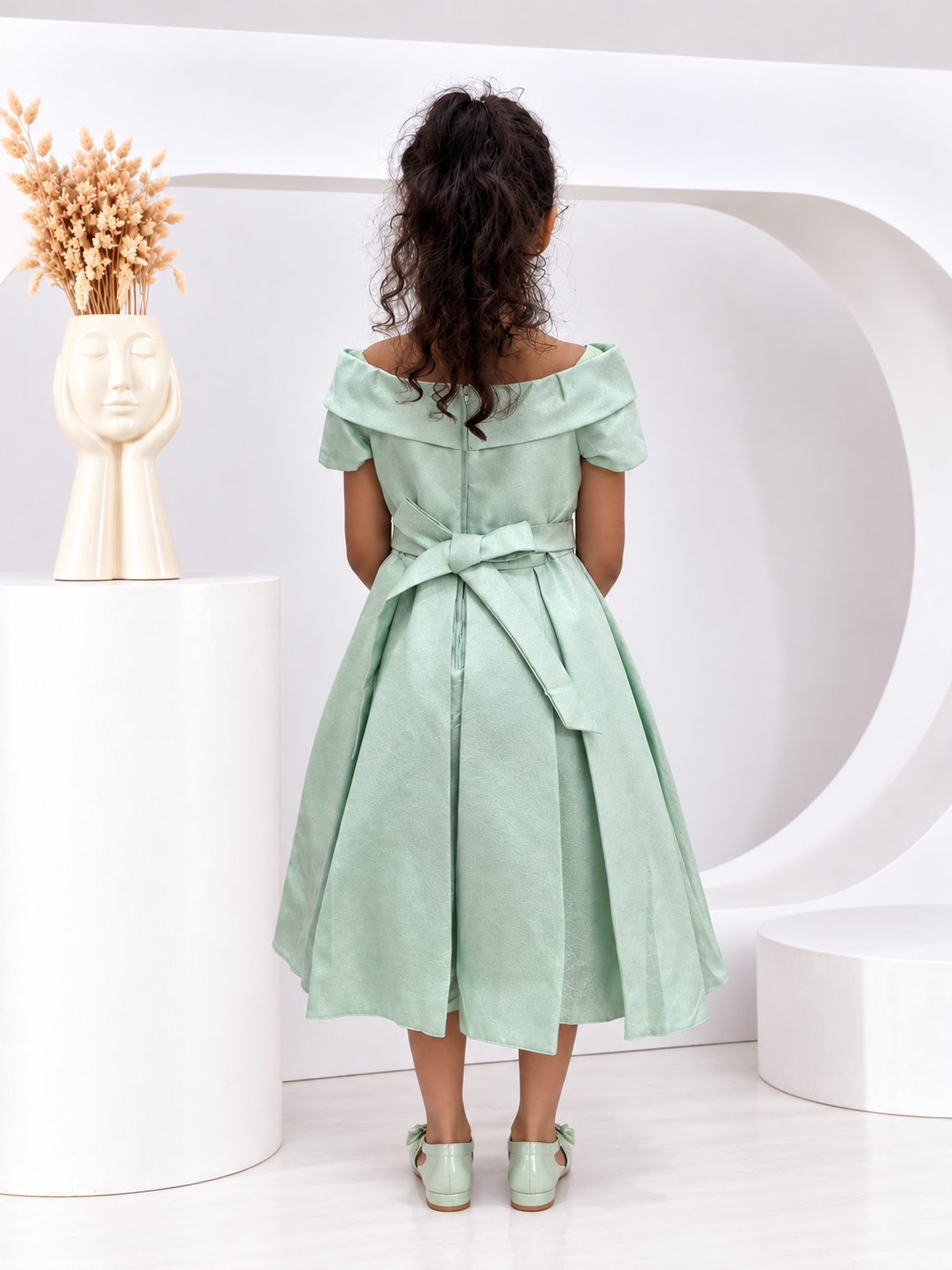 A young girl viewed from behind wearing a mint green off-the-shoulder midi dress with a matching waist tie, standing in a modern white studio setting.