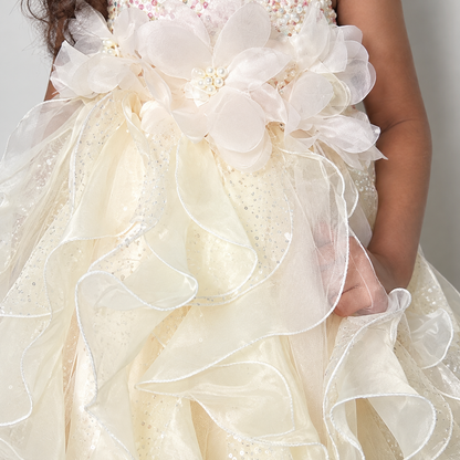 Close-up of a child wearing a formal cream or ivory dress with layered sheer organza ruffles, large fabric flower appliqués, and pearl-like bead accents.