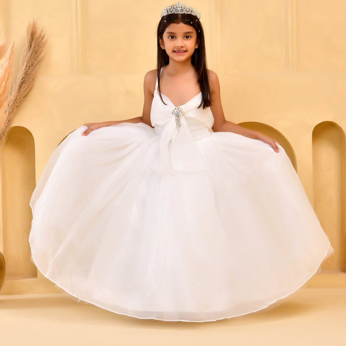 A young girl wearing a white ballgown with a tiara, sitting on a beige platform in front of a matching beige background.