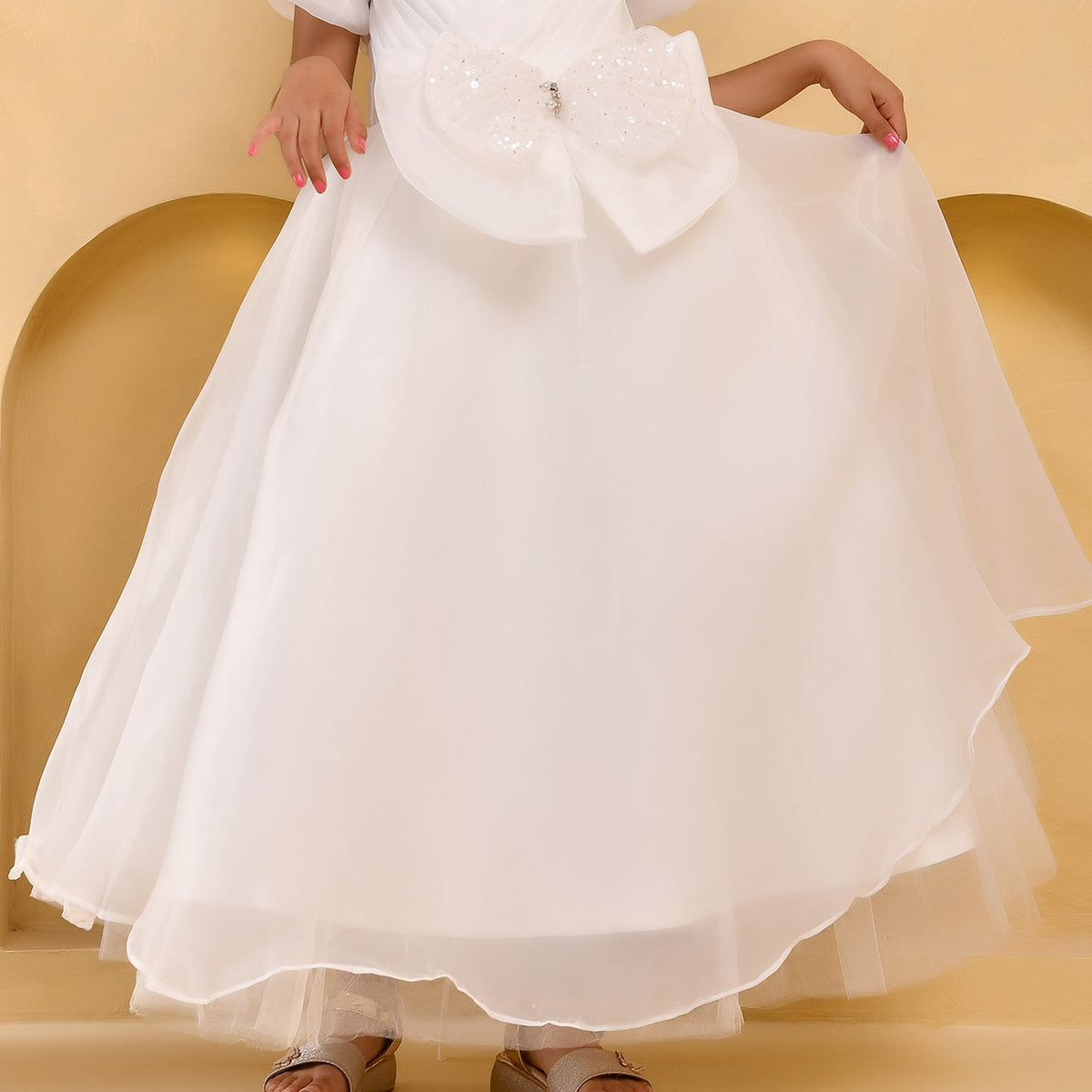 A child wearing a voluminous white tulle dress with a large embellished bow at the waist, standing against a warm beige arched backdrop.