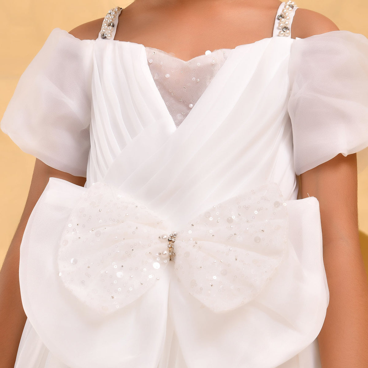 A close-up of a white formal dress with sheer puff sleeves, a V-neckline, and a large bow detail on the front covered in silver sequins and embellishments.