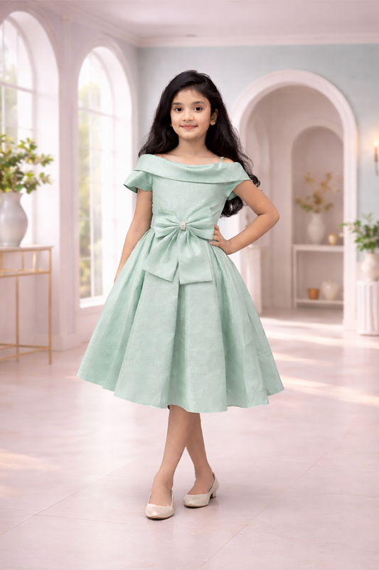 A young girl in a light green dress from Nibblekins with a bow tie standing in an elegant room.