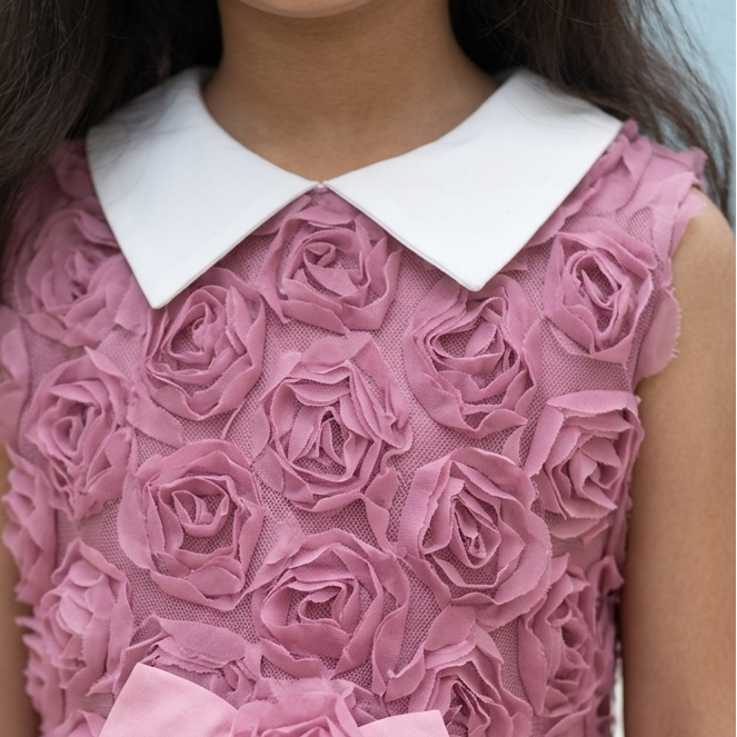 A close-up of a young girl wearing a sleeveless dusty pink dress with a 3D rose texture, a white Peter Pan collar, and a matching pink fabric bow at the waist.
