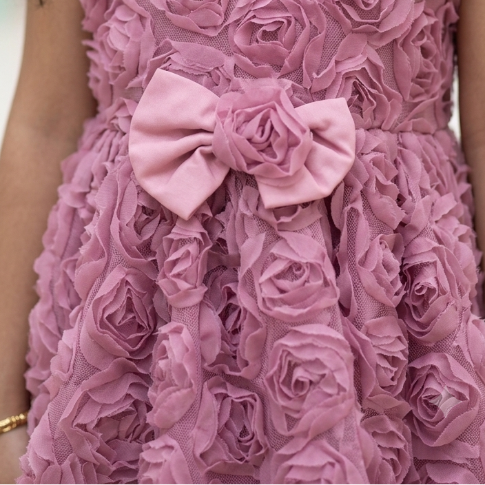A close-up of a young girl wearing a sleeveless dusty pink dress with a 3D rose texture, a white Peter Pan collar, and a matching pink fabric bow at the waist.