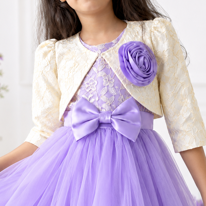 A young girl wearing a formal lavender tulle dress with a matching lavender bow at the waist, a cream lace bodice, and a cream textured short jacket with a large lavender fabric flower on the chest, standing in a bright indoor setting.