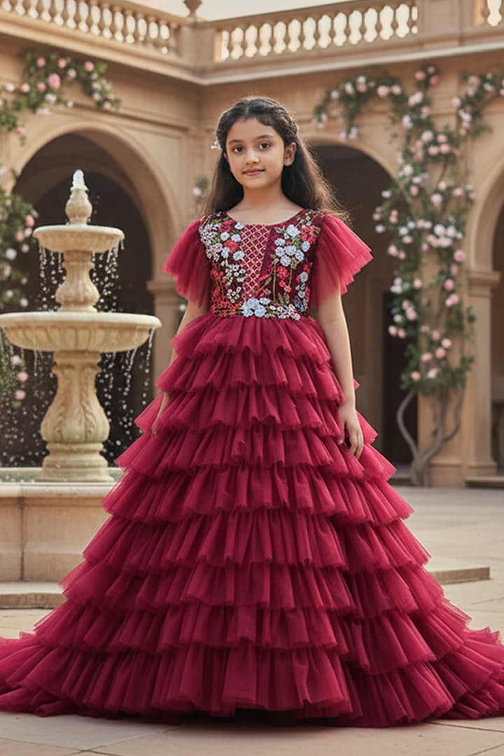 A Young girl in a red gown with floral details from Nibblekins standing in front of a decorative fountain.