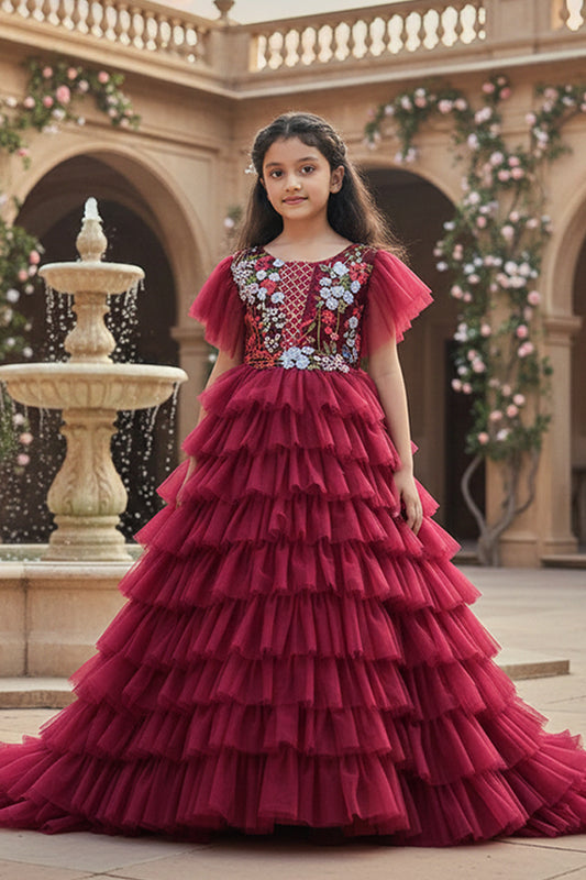 A Young girl in a red gown with floral details from Nibblekins standing in front of a decorative fountain.
