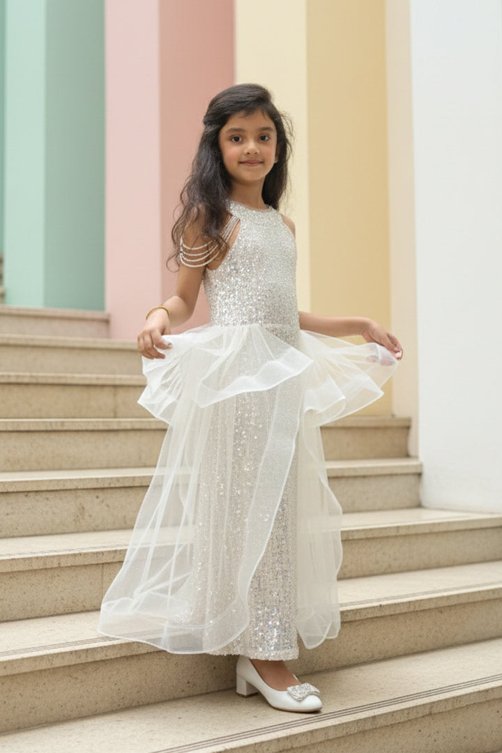A Young girl in a white sequin dress from Nibblekins is standing on a set of stairs with pastel-colored walls.