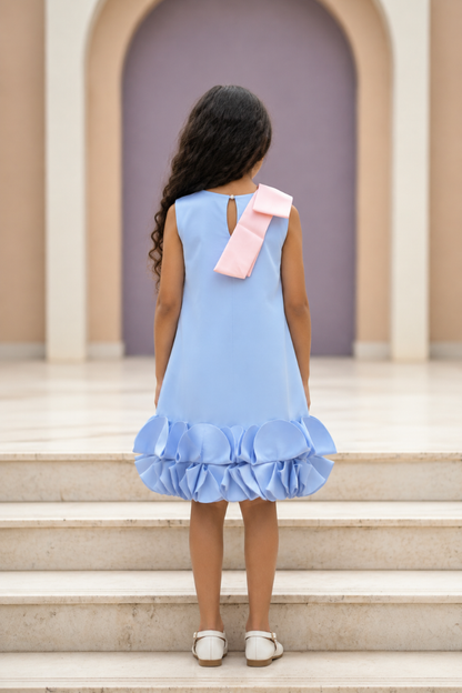 A young girl viewed from behind wearing a sleeveless light blue dress with a large pink bow on the back and a ruffled hem, standing on stone steps in front of an arched doorway.