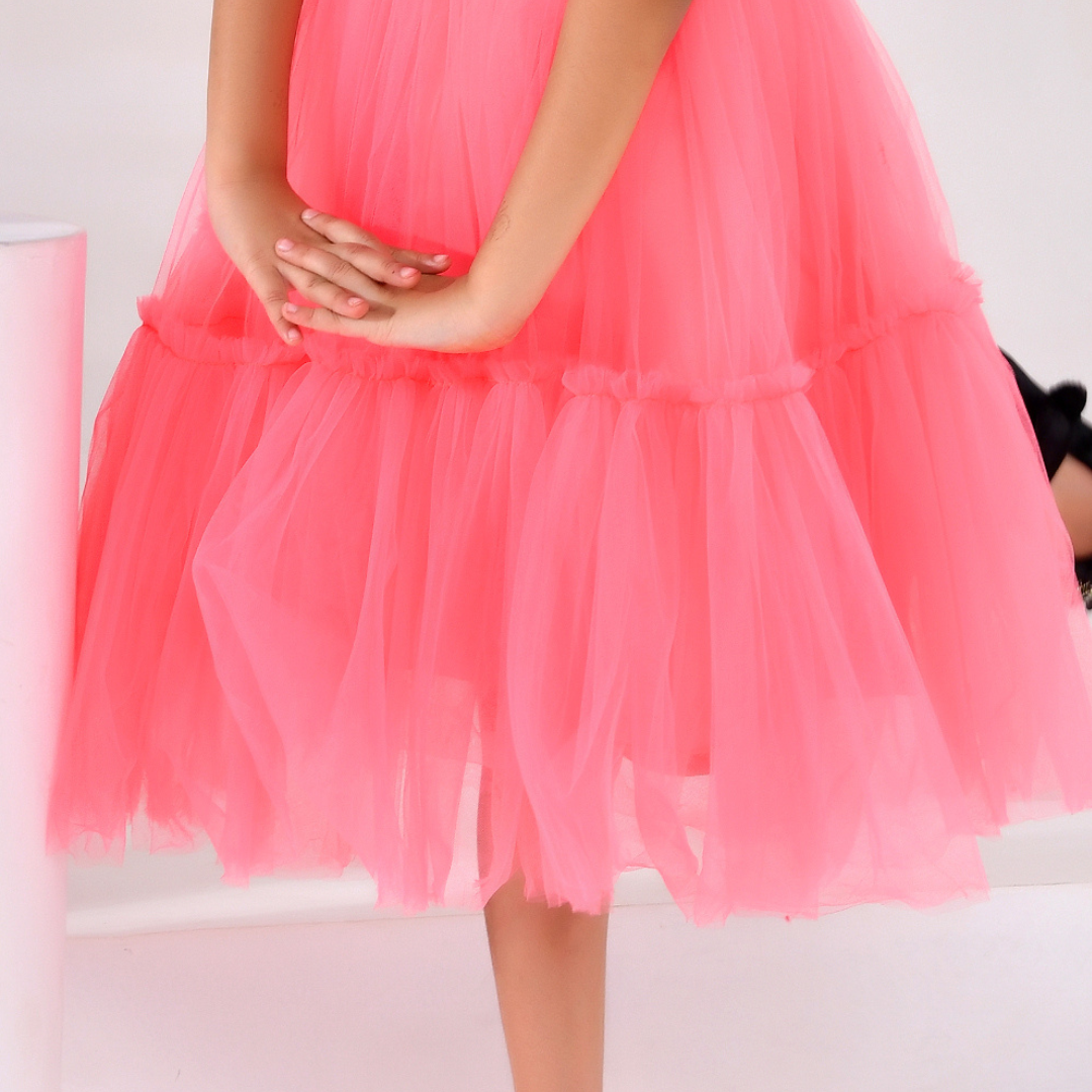 A close-up of a child wearing a bright pink tiered tulle dress, showing the layered mesh skirt and hands clasped in front.