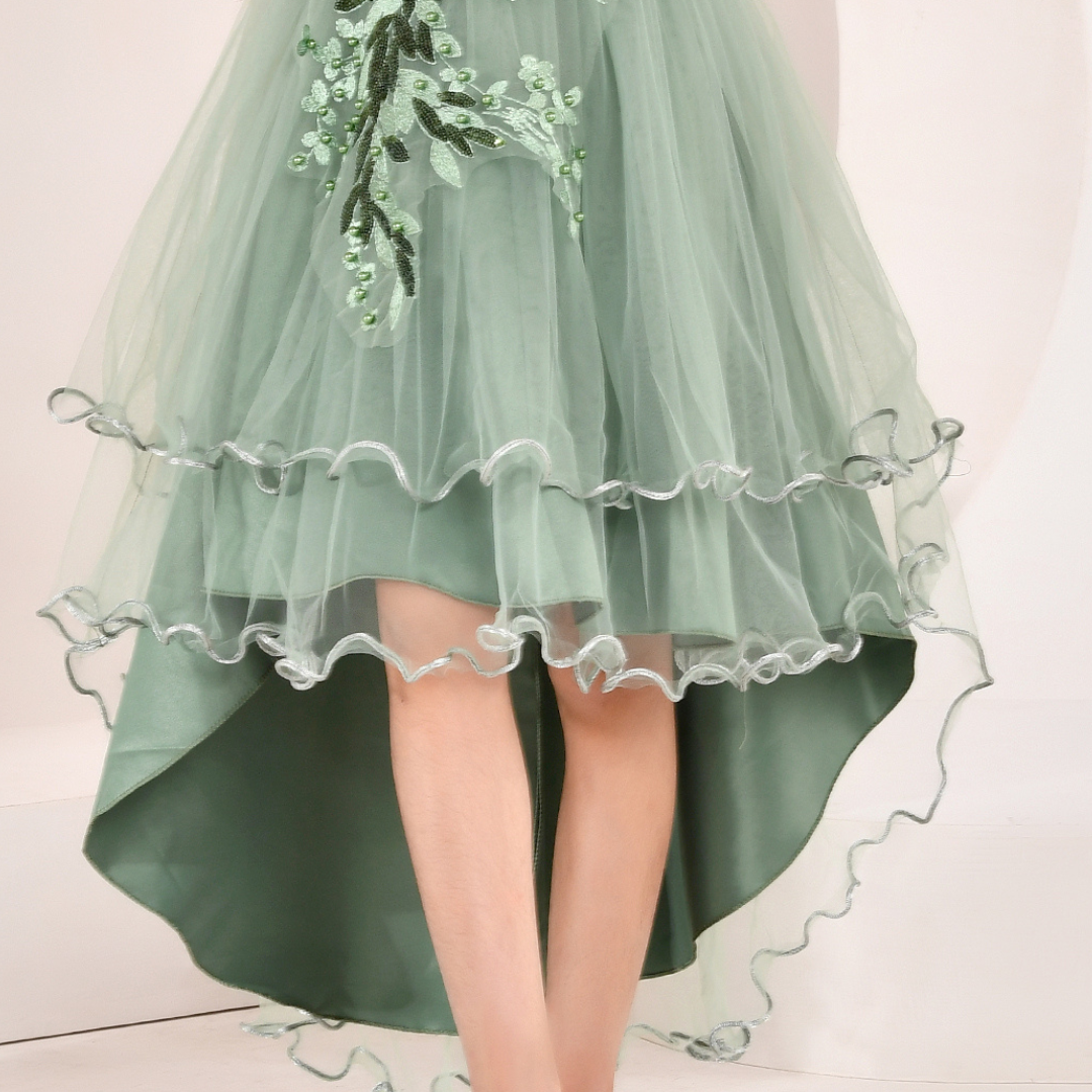 A close-up of a model wearing a sage green high-low tulle dress with floral embroidery on the bodice and layered ruffled hems.