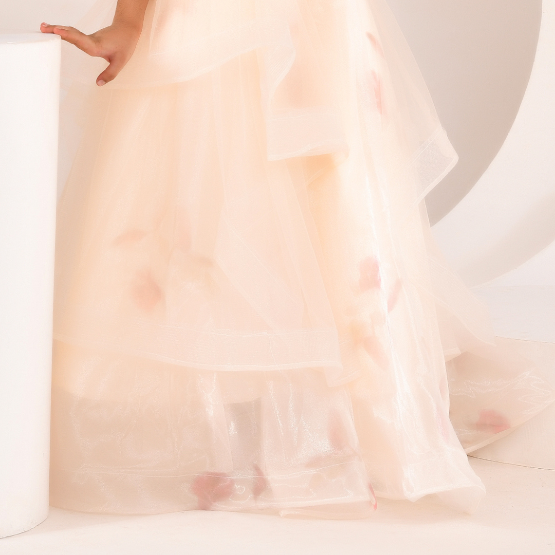 A close-up of a sheer, layered tulle dress or gown in a very light blush pink, with soft pink floral accents scattered through the fabric. A hand is visible resting on a white cylindrical pedestal.