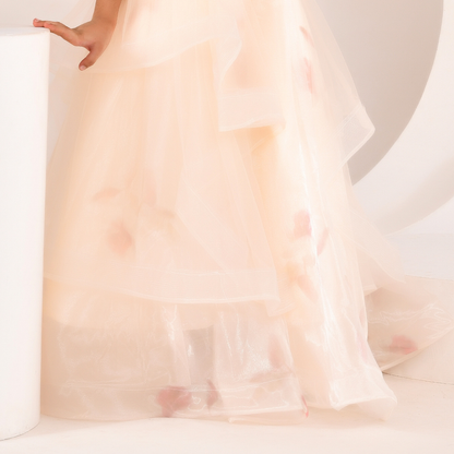 A close-up of a sheer, layered tulle dress or gown in a very light blush pink, with soft pink floral accents scattered through the fabric. A hand is visible resting on a white cylindrical pedestal.