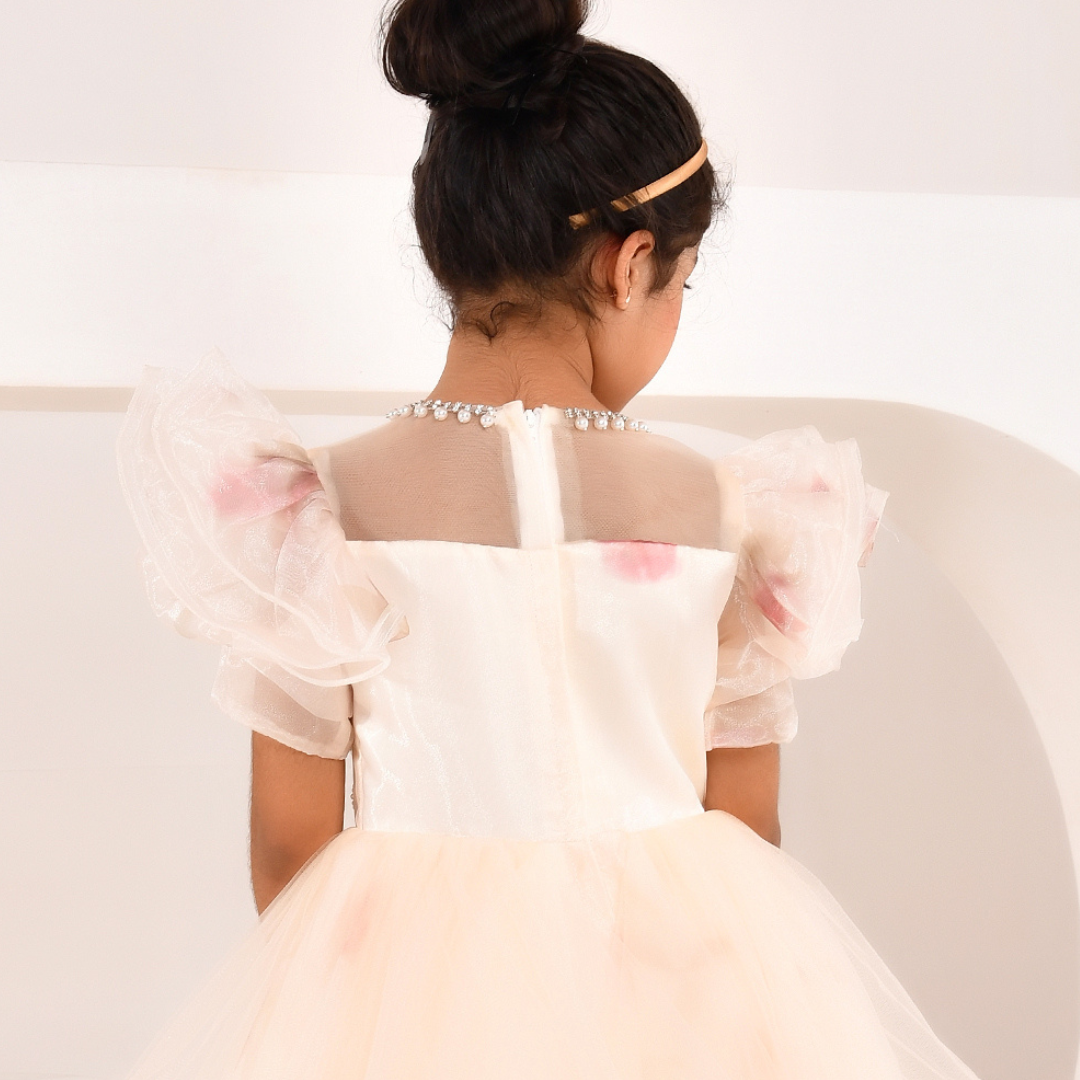 A young girl viewed from behind wearing a formal party dress with sheer puff sleeves, a pearl-embellished neckline, and a light pink tulle skirt, set against a clean white background.