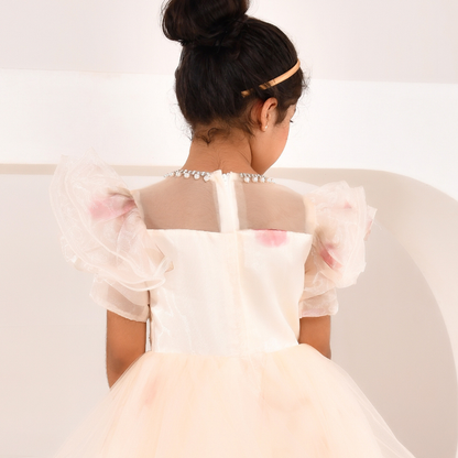 A young girl viewed from behind wearing a formal party dress with sheer puff sleeves, a pearl-embellished neckline, and a light pink tulle skirt, set against a clean white background.