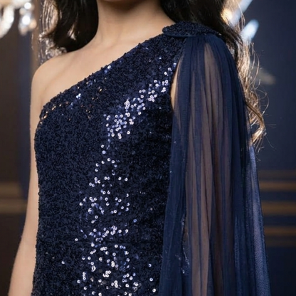 A close-up of a woman wearing a navy blue one-shoulder sequined gown with a sheer draped sleeve.