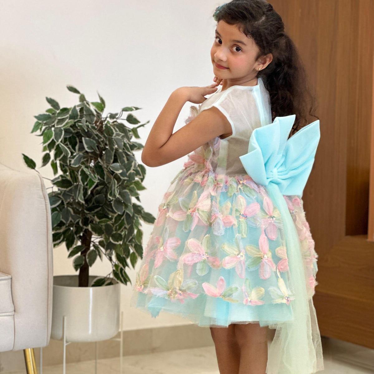 A young girl modeling a light blue and pink tulle dress with a large bow on the back, standing barefoot in an indoor setting.