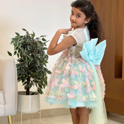 A young girl modeling a light blue and pink tulle dress with a large bow on the back, standing barefoot in an indoor setting.