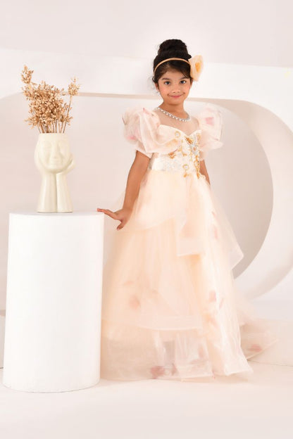 Young girl in a pink dress standing in a softly lit room with a vase of dried flowers in the background.