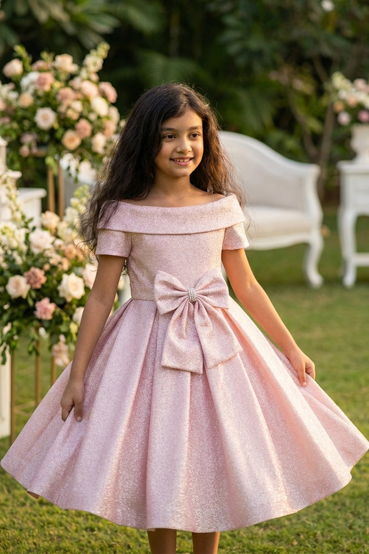 Young girl in a pink off-shoulder dress with a bow standing outdoors.